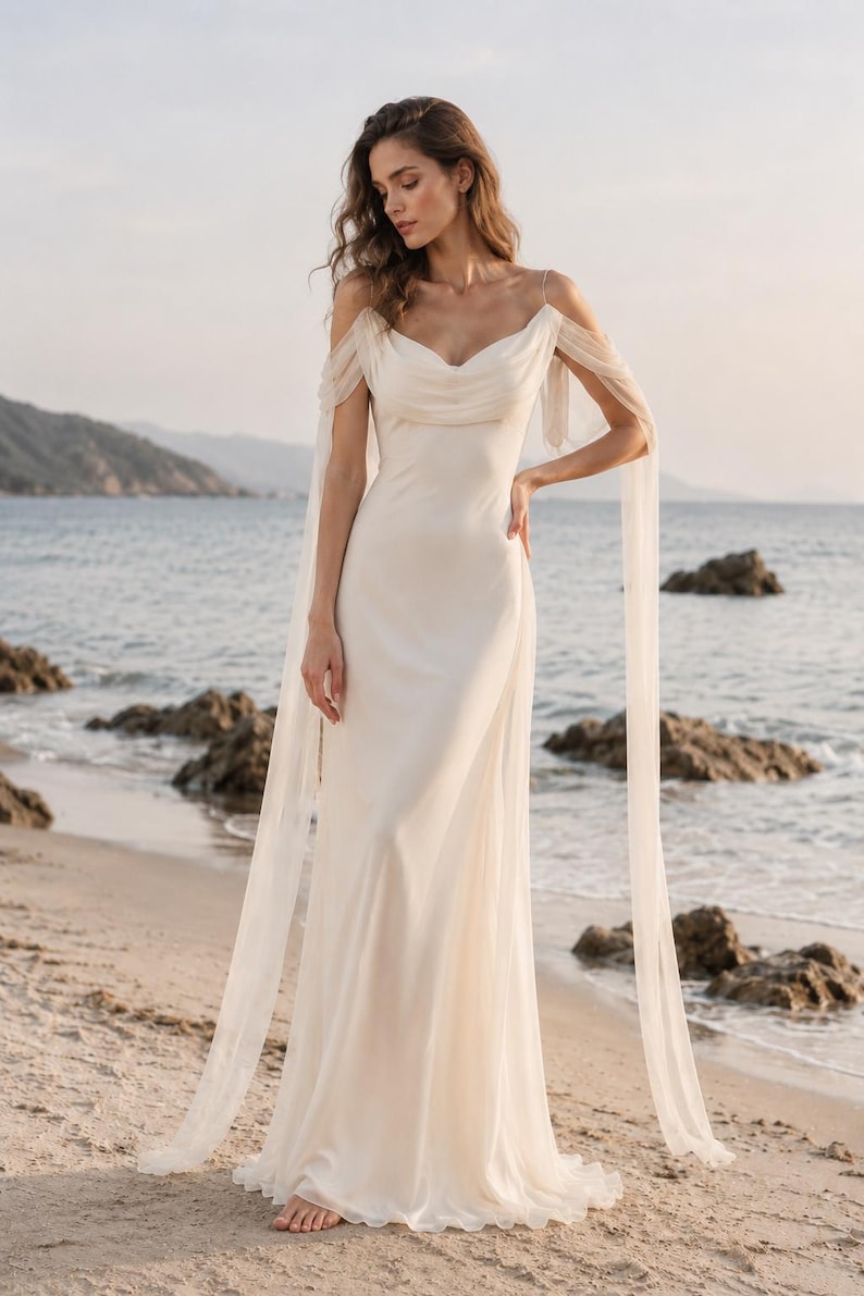 May include: A floor-length ivory gown featuring a draped cowl neckline, thin spaghetti straps, and sheer off-the-shoulder sleeves that extend into long, flowing fabric panels. The dress has a fitted bodice and a smooth, floor-length skirt.