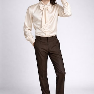 May include: Cream-colored satin shirt with a large bow tie, paired with dark brown trousers and black leather loafers. The outfit is a classic, elegant style.