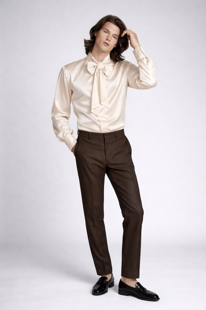 May include: Cream-colored satin shirt with a large bow tie, paired with dark brown trousers and black leather loafers. The outfit is a classic, elegant style.