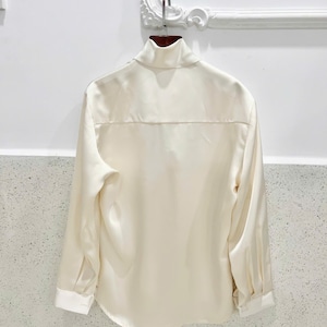 May include: Cream-colored long-sleeve shirt with a high collar and white cuffs. The back of the shirt is visible, hanging on a wooden hanger. The shirt has a smooth, silky texture and a classic design.