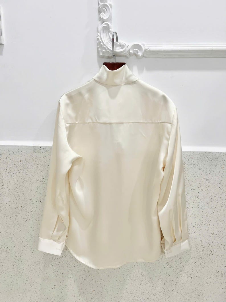 May include: Cream-colored long-sleeve shirt with a high collar and white cuffs. The back of the shirt is visible, hanging on a wooden hanger. The shirt has a smooth, silky texture and a classic design.