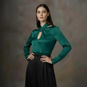 Puede incluir: Blusa de sat&eacute;n verde esmeralda con mangas largas y cuello con abertura. La blusa presenta una botonadura frontal y se combina con pantalones negros de pernera ancha. El conjunto se presenta sobre un fondo neutro.