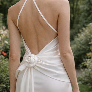 May include: Ivory-colored wedding dress with a halter neckline and a low back. The dress features a rose detail at the waist and a flowing skirt. The dress is made of a smooth, silky fabric, perfect for a wedding or special occasion.