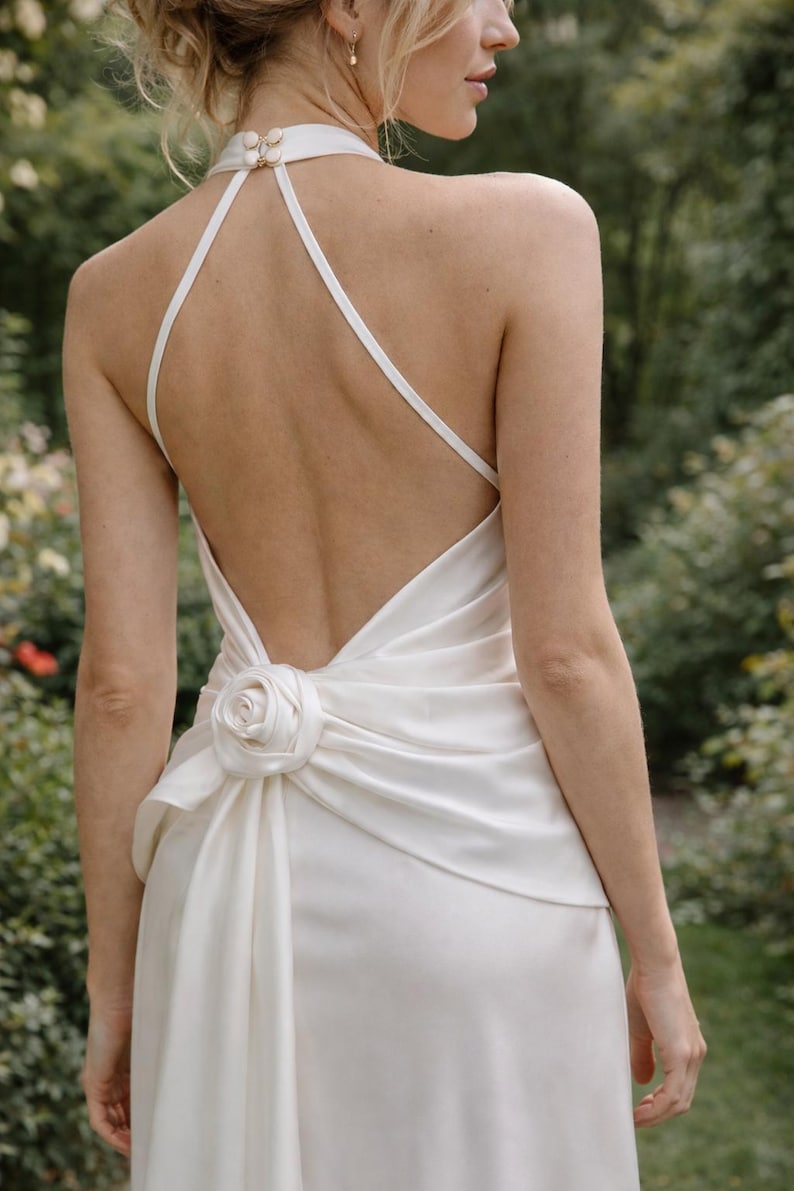 May include: Ivory-colored wedding dress with a halter neckline and a low back. The dress features a rose detail at the waist and a flowing skirt. The dress is made of a smooth, silky fabric, perfect for a wedding or special occasion.