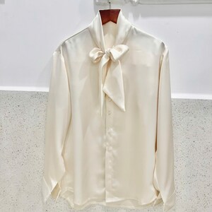 May include: Ivory-colored long-sleeve blouse with a bow tie collar. The blouse is made of a smooth, silky material and features button closures down the front and at the cuffs. The garment is hanging on a wooden hanger.
