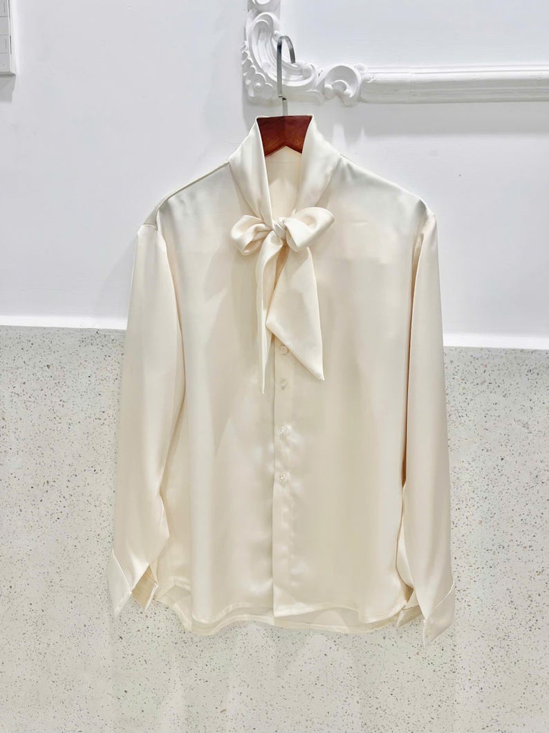 May include: Ivory-colored long-sleeve blouse with a bow tie collar. The blouse is made of a smooth, silky material and features button closures down the front and at the cuffs. The garment is hanging on a wooden hanger.