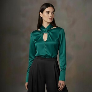 Puede incluir: Blusa de sat&eacute;n verde esmeralda con mangas largas y cuello con abertura. La blusa tiene una botonadura frontal y se combina con pantalones negros de pernera ancha. La tela tiene un brillo sutil, lo que sugiere un estilo formal o elegante.