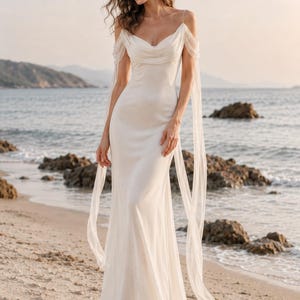 May include: A floor-length white gown featuring a draped cowl neckline, thin spaghetti straps, and off-the-shoulder sheer fabric sleeves that extend into long, flowing panels reaching toward the hemline, creating a soft, fluid silhouette.