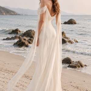 May include: A floor-length off-white gown featuring a draped bodice, thin spaghetti straps, and long, flowing sheer fabric sleeves that extend from the shoulders. The dress has a low back design and a full-length skirt made of lightweight material.