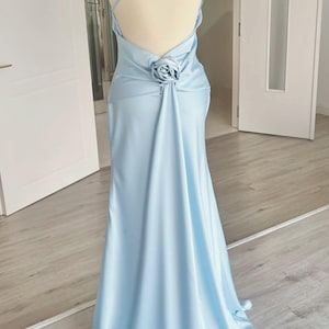 May include: A light blue, floor-length gown with a halter neckline and a low back. The dress features a rose detail at the waist and a flowing skirt. The dress is displayed on a mannequin in a well-lit room.