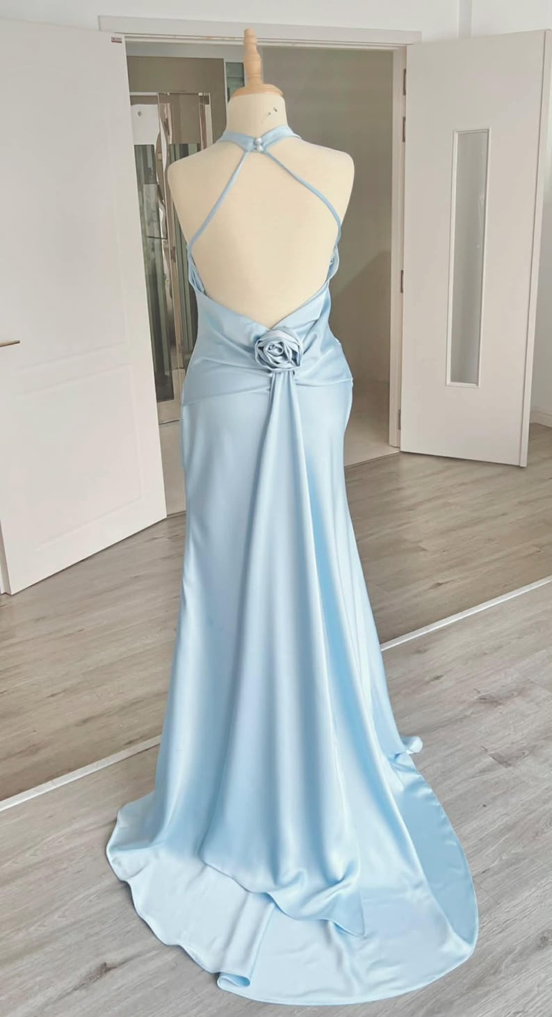 May include: A light blue, floor-length gown with a halter neckline and a low back. The dress features a rose detail at the waist and a flowing skirt. The dress is displayed on a mannequin in a well-lit room.