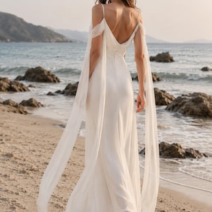 May include: A floor-length white gown featuring a deep V-back, thin spaghetti straps, and long, flowing off-the-shoulder sheer sleeves that drape down to the hem. The dress has a fitted bodice and a skirt that falls in a straight, fluid silhouette.