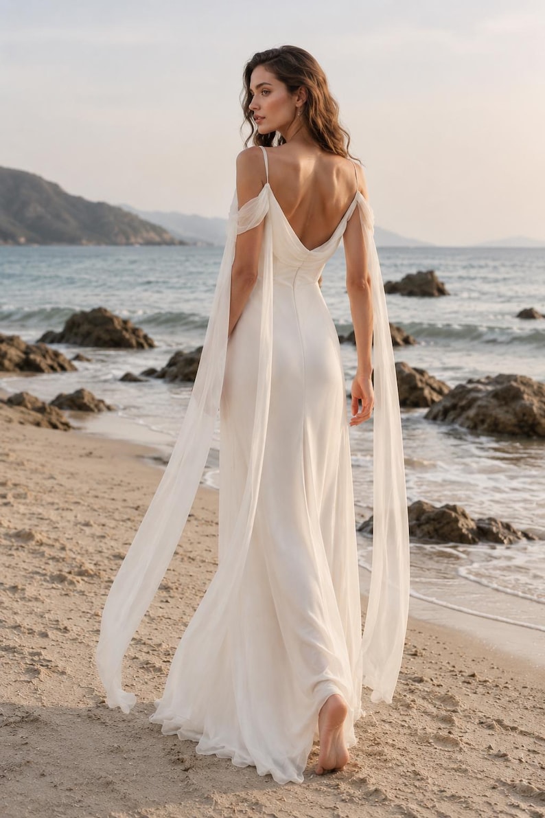 May include: A floor-length white gown featuring a deep V-back, thin spaghetti straps, and long, flowing off-the-shoulder sheer sleeves that drape down to the hem. The dress has a fitted bodice and a skirt that falls in a straight, fluid silhouette.
