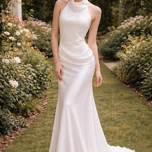 May include: A white, floor-length gown with a halter neckline and a flowing skirt. The dress is made of a smooth, silky fabric and features a slight train. The model is standing in a garden setting with greenery and flowers.