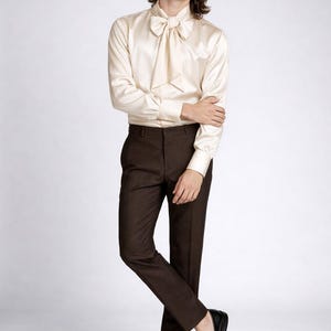 May include: A cream-colored satin shirt with a large bow tie, paired with dark brown trousers and black leather loafers. The outfit is a classic, elegant style, suitable for formal occasions. The model is standing in a studio setting.