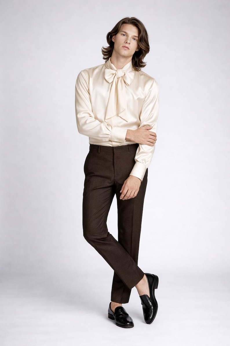 May include: A cream-colored satin shirt with a large bow tie, paired with dark brown trousers and black leather loafers. The outfit is a classic, elegant style, suitable for formal occasions. The model is standing in a studio setting.