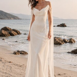 May include: A floor-length ivory gown featuring a draped cowl neckline, thin spaghetti straps, and long, sheer, flowing sleeves that extend from the shoulders. The garment is made of a smooth, lightweight fabric that falls in a straight silhouette.