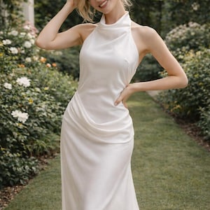 May include: A white halter neck wedding dress with a draped bodice and a flowing skirt. The dress is made of a smooth, silky fabric. The model is smiling and standing in a garden setting with green foliage.