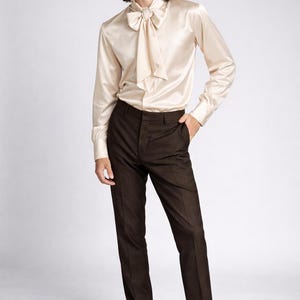 May include: A cream-colored satin shirt with a bow tie, paired with dark brown trousers and black leather loafers. The long-sleeved shirt has a flowing bow detail at the neck. The trousers are tailored and the loafers are polished.