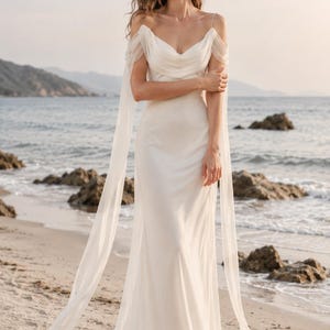 May include: A floor-length ivory gown featuring a draped cowl neckline, thin spaghetti straps, and long, sheer, flowing off-the-shoulder sleeves that extend to the floor. The dress has a fitted bodice and a smooth, flowing skirt silhouette.
