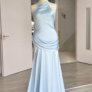 May include: A light blue satin gown displayed on a mannequin. The dress features a halter neckline, a draped bodice, and a flowing skirt. The gown is floor-length and has a subtle sheen.