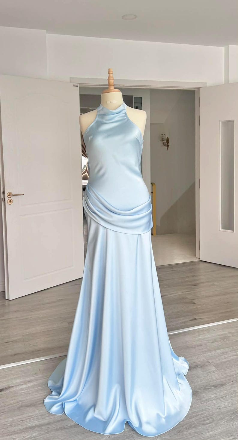 May include: A light blue satin gown displayed on a mannequin. The dress features a halter neckline, a draped bodice, and a flowing skirt. The gown is floor-length and has a subtle sheen.