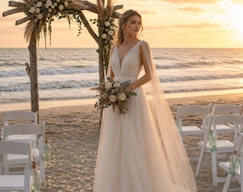 May include: A floor-length ivory wedding dress with a deep V-neckline, sheer cape sleeves, and a flowing tulle skirt. The dress is paired with a bouquet of dried flowers and pampas grass, standing before a driftwood arch decorated with white roses.