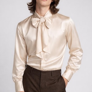 May include: A cream-colored satin shirt with a large bow tie and long sleeves. The shirt is paired with brown trousers. The fabric has a slight sheen, and the overall look is elegant and formal.