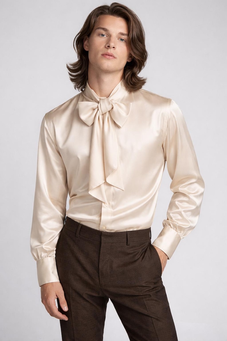 May include: A cream-colored satin shirt with a large bow tie and long sleeves. The shirt is paired with brown trousers. The fabric has a slight sheen, and the overall look is elegant and formal.