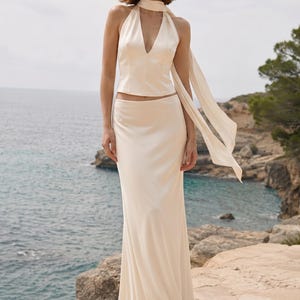 May include: A two-piece set consisting of a sleeveless, cream-colored satin halter top with a deep V-neckline and a matching long, floor-length satin skirt. The top features an integrated scarf-like neck tie that drapes down the side.