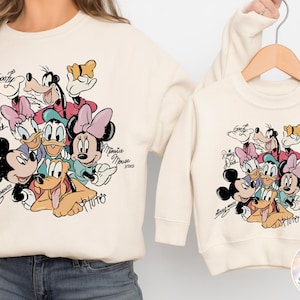 May include: Cream-colored sweatshirts featuring a colorful graphic of Disney characters including Mickey Mouse, Minnie Mouse, Donald Duck, Daisy Duck, Goofy, and Pluto. The characters' names are written in cursive. A smaller version of the sweatshirt is on a hanger.
