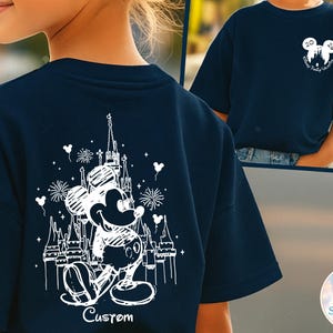 May include: Navy blue t-shirt featuring a white graphic of Mickey Mouse in front of a castle, with fireworks and the word "Custom" below. The shirt also has a small Mickey Mouse head logo with text on the front.