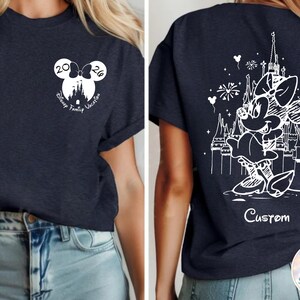 May include: Navy blue t-shirt featuring a white Minnie Mouse design on the back with a castle and fireworks. The front has a Mickey Mouse head with the text "Disney Family Vacation 2026".