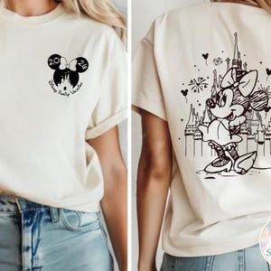 May include: Cream-colored t-shirt with black graphics. The front features a Mickey Mouse ear design with the numbers "20" and "26" and the words "Disney Family Vacation". The back displays a Minnie Mouse illustration in front of a castle.