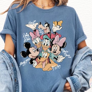 May include: A blue t-shirt featuring a graphic of Disney characters including Mickey Mouse, Minnie Mouse, Donald Duck, Daisy Duck, Goofy, and Pluto. The names of the characters are written in cursive script around the image.