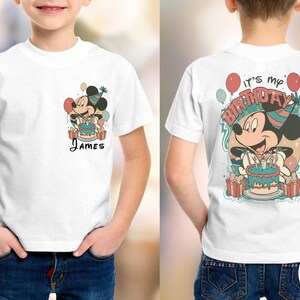 May include: White t-shirt featuring a cartoon Mickey Mouse design with a birthday cake and balloons. The front of the shirt has the name "James". The back of the shirt says "It's my Birthday".