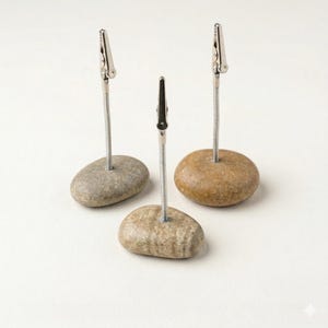 Pode incluir: Três porta-cartões de pedra natural. Cada suporte apresenta uma presilha de metal prateado presa a uma mola helicoidal, montada em uma pedra lisa e arredondada. As pedras variam em cor, do cinza claro ao marrom.