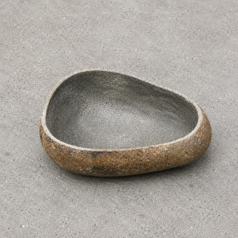 May include: A bowl with an irregular, organic shape resembling a river stone. The exterior has a textured, mottled brown and tan finish, while the interior features a smooth, speckled dark gray surface with a deep, concave basin design.