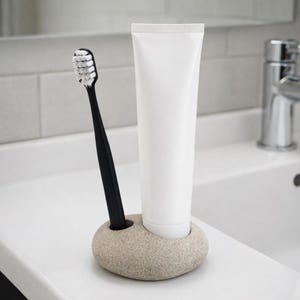 May include: A black toothbrush with white bristles and a white tube of toothpaste are in a beige stone holder. The holder is on a white countertop, with a white tiled wall and a chrome faucet in the background. The toothbrush and toothpaste are for oral hygiene.