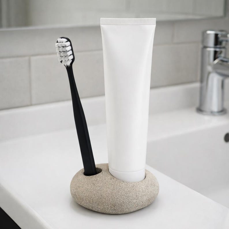 May include: A black toothbrush with white bristles and a white tube of toothpaste are in a beige stone holder. The holder is on a white countertop, with a white tiled wall and a chrome faucet in the background. The toothbrush and toothpaste are for oral hygiene.