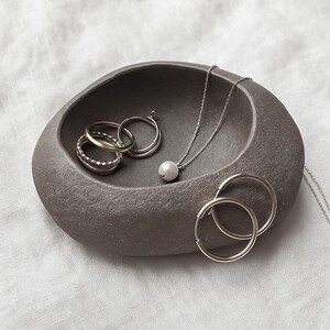 May include: A grey, stone-effect jewellery dish contains silver rings, a pearl pendant necklace, and a pair of hoop earrings. The jewellery is arranged on a white, textured surface, creating a minimalist aesthetic. The dish has a smooth, organic shape.
