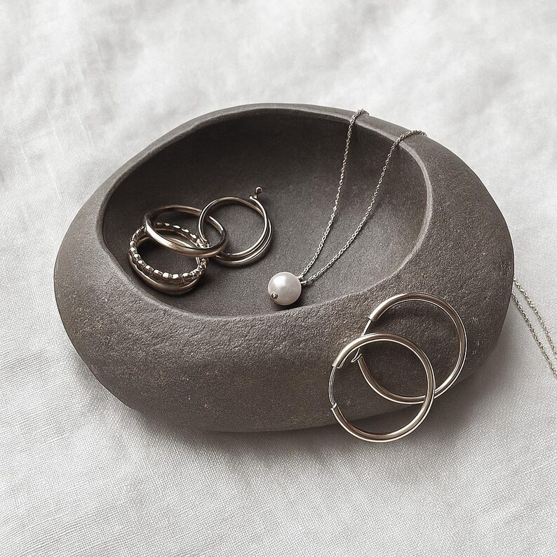 May include: A grey, stone-effect jewellery dish contains silver rings, a pearl pendant necklace, and a pair of hoop earrings. The jewellery is arranged on a white, textured surface, creating a minimalist aesthetic. The dish has a smooth, organic shape.