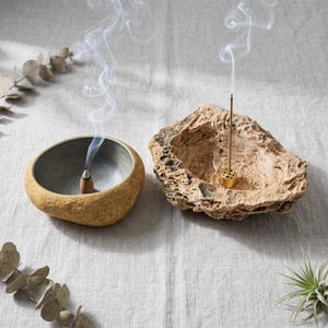 May include: A smooth, rounded stone incense burner with a grey interior and a burning cone, alongside a porous, textured rock incense holder with a gold-toned metal stand holding a thin, upright burning incense stick with rising smoke.