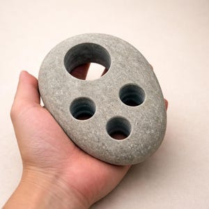 May include: A gray, oval-shaped stone with three small holes and one large hole. The stone is held in a hand, showcasing its natural texture and design. This unique item could be used as a toothbrush holder or a decorative piece.