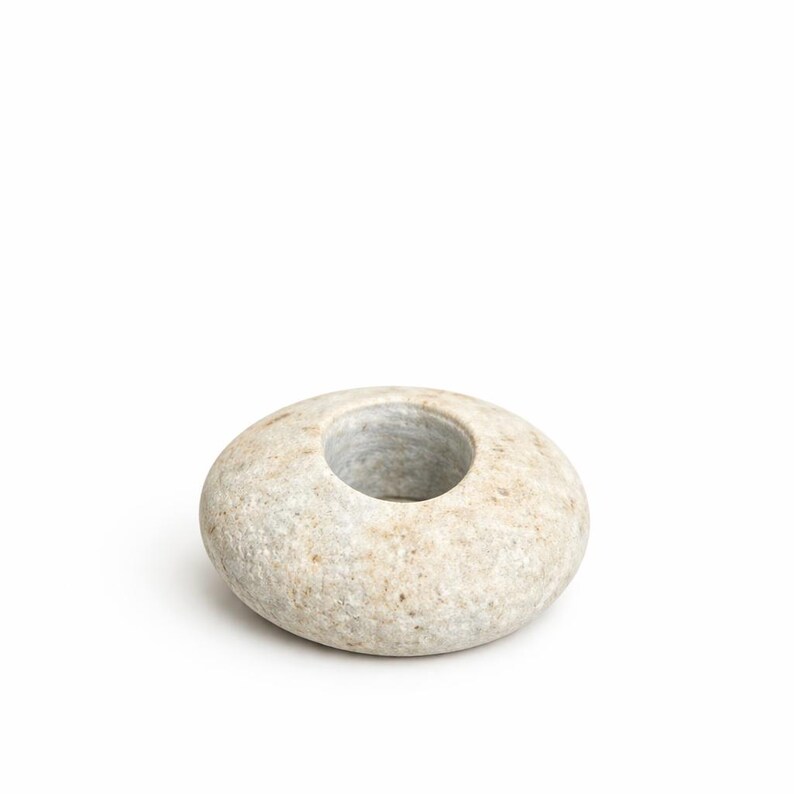 May include: A smooth, oval-shaped stone candle holder with a central circular opening. The stone has a light beige color with subtle variations in tone, and a natural, textured surface. The candle holder is designed to hold a single candle.