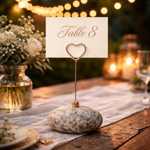 May include: A wedding table number holder with a heart-shaped wire and a stone base. The card reads "Table 8" in elegant script. The setting includes flowers, candles, and a rustic wooden table, creating a romantic ambiance.