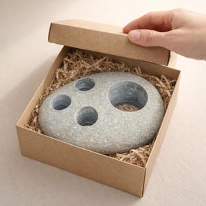 May include: A gray stone toothbrush holder with four circular openings, presented in a brown cardboard box with shredded paper filler. The holder has a natural, textured appearance, suggesting a handcrafted item.
