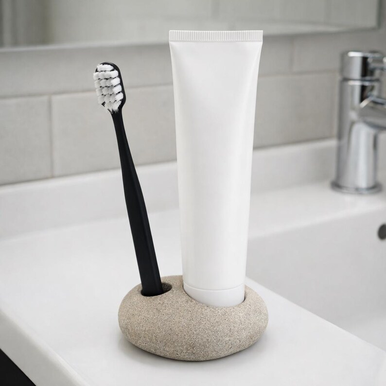 Puede incluir: Un cepillo de dientes negro con cerdas blancas y un tubo de pasta de dientes blanco est&aacute;n en un soporte de piedra beige. El soporte est&aacute; sobre una encimera blanca, con una pared de azulejos blancos y un grifo cromado en el fondo. Para la higiene bucal.