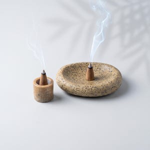 May include: A cylindrical stone incense holder and a shallow, oval-shaped stone incense dish, both featuring a speckled beige and brown texture. Each piece holds a burning brown incense cone, with thin wisps of white smoke rising upward.