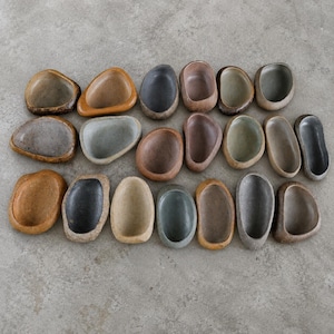 Peut inclure: Une collection de vingt petits bols en céramique de forme ovale disposés sur trois rangées. Chaque bol présente une texture organique unique rappelant la pierre, avec des émaux aux tons terreux, bruns, gris, beiges et anthracite, et un intérieur lisse.